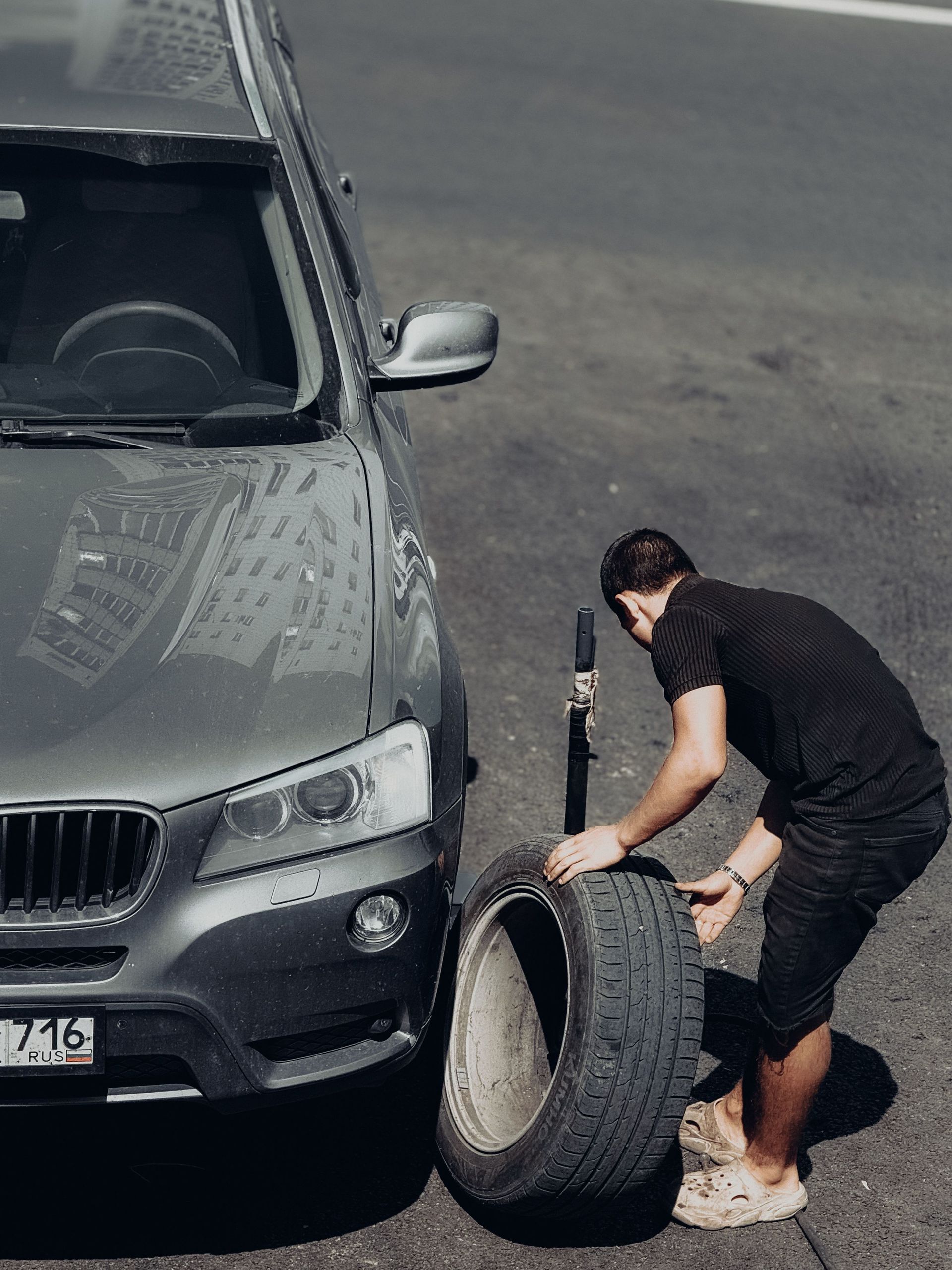 A young man changes a car tire on an outdoor road under the bright sunlight, highlighting the DIY task.
